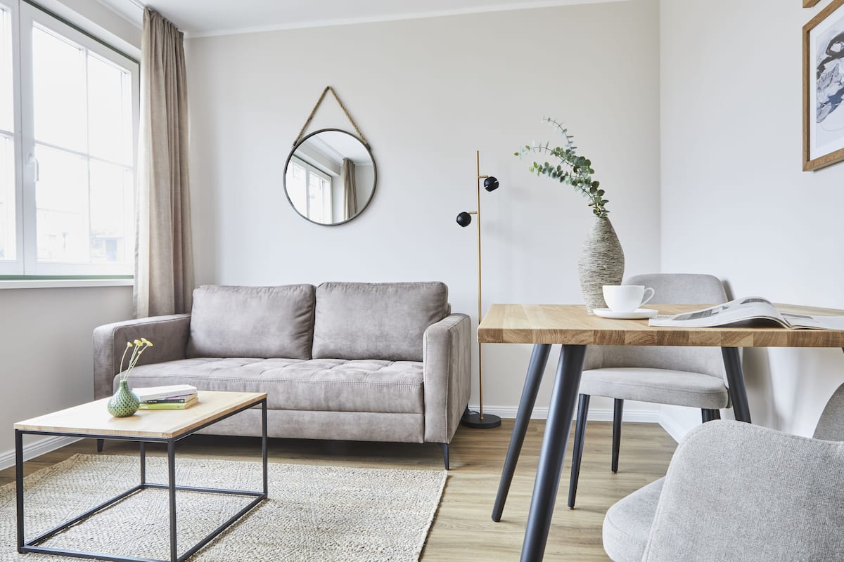A modern living room with a grey sofa, a wooden coffee table, a small side table with a vase, a floor lamp, and a dining table with chairs.