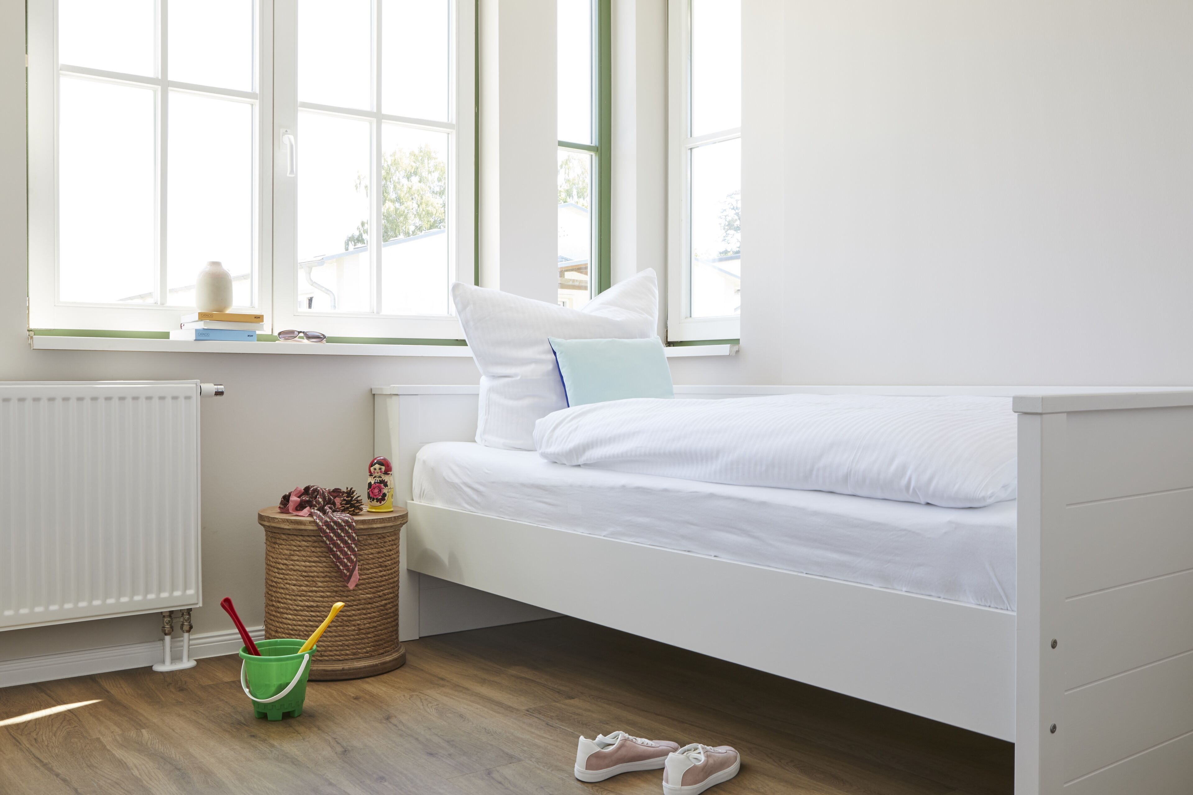 Une chambre avec un lit blanc, une fenêtre donnant sur un paysage, un radiateur, une petite poubelle et une paire de chaussures posées sur le sol.