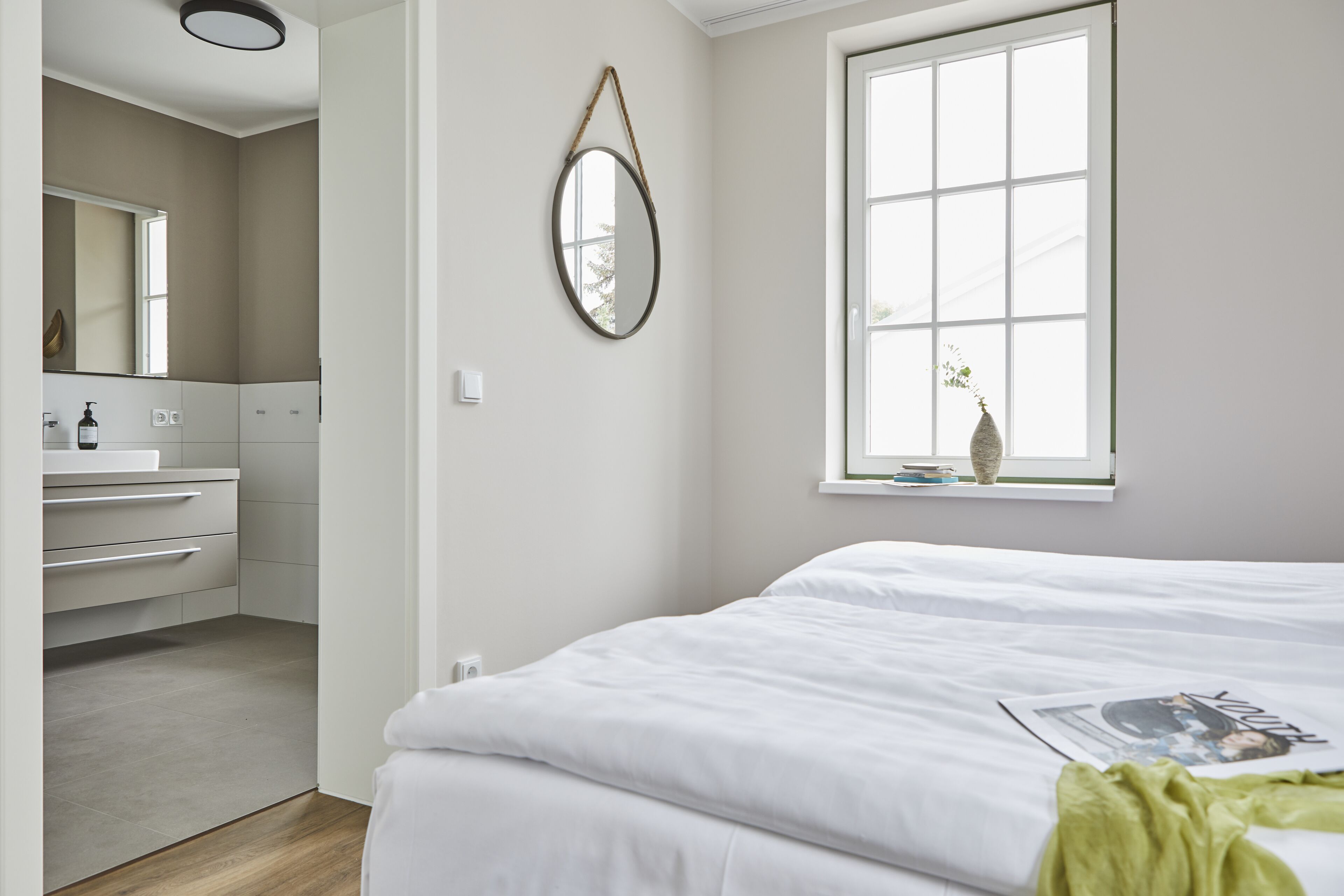 A bedroom with a large window, a bed, a bedside table, and a mirror.