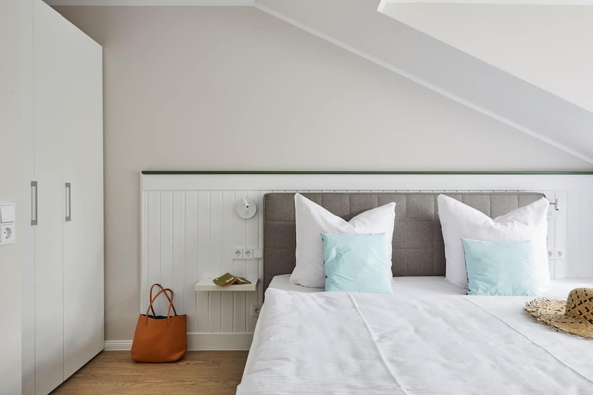 A bedroom with a sloped ceiling, a bed with white bedding, two light blue pillows, a brown handbag, and a straw hat on the bed.