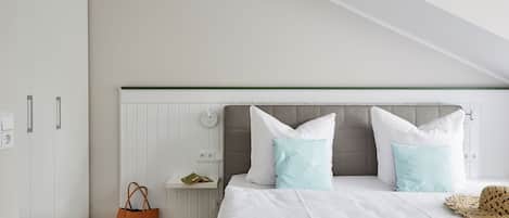 A bedroom with a sloped ceiling, a bed with white bedding, two light blue pillows, a brown handbag, and a straw hat on the bed.