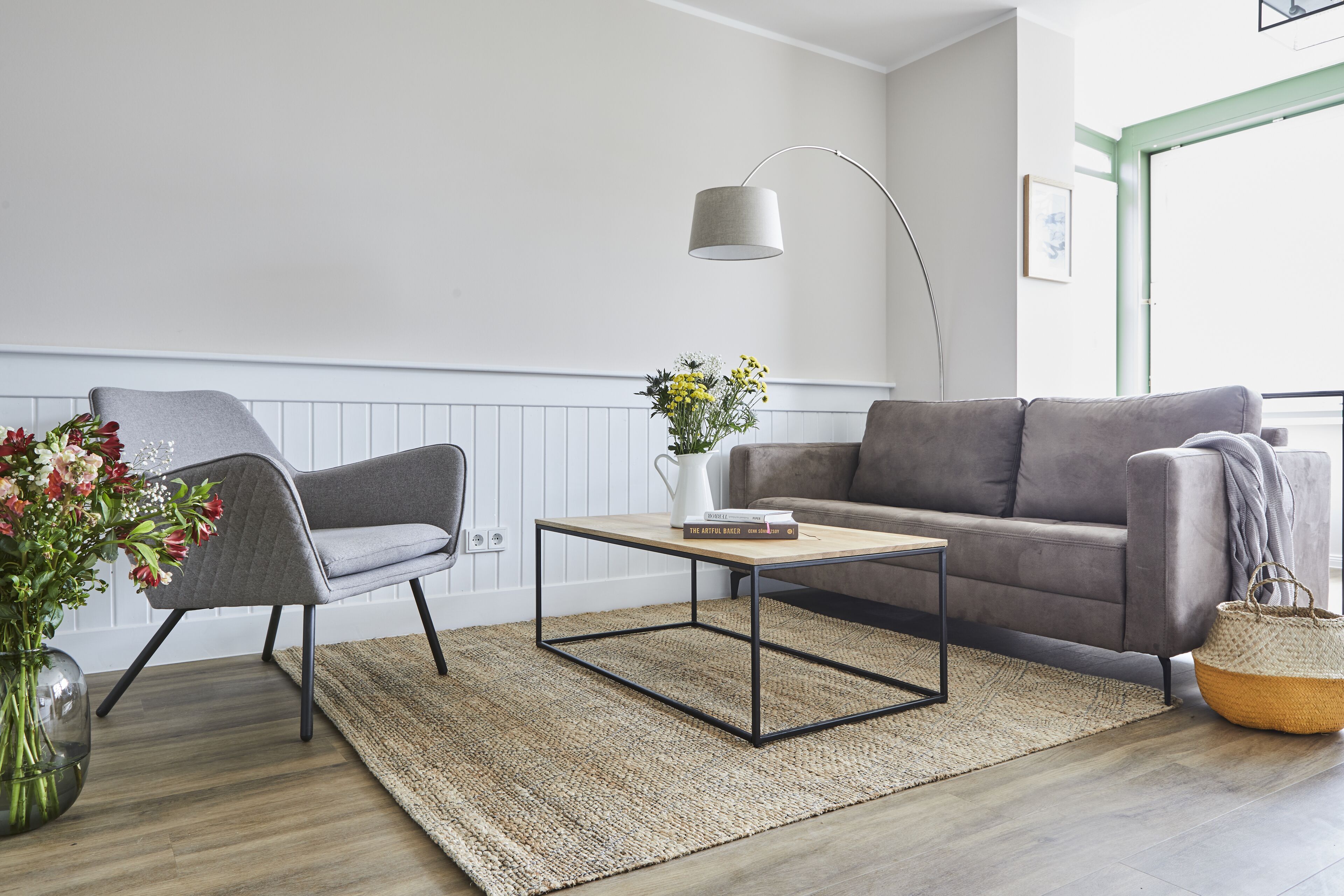 A modern living room with a grey sofa, a grey armchair, a wooden coffee table, a floor lamp, and a vase with flowers.