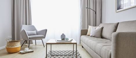 Ein modernes Wohnzimmer mit einer Couch, einem Sessel, einem Couchtisch und einem großen Fenster mit Vorhängen.