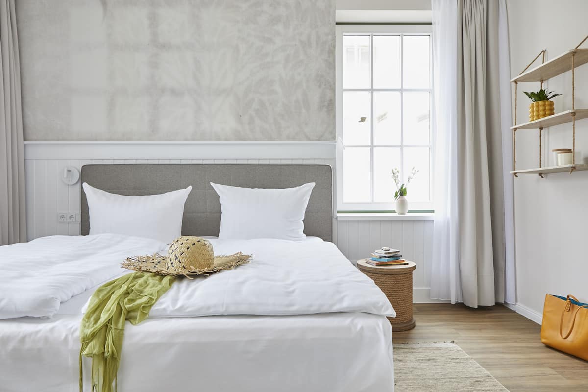 A neatly made bed with white linens and a straw hat on it. There is a small round table with books and a potted plant beside the bed.
