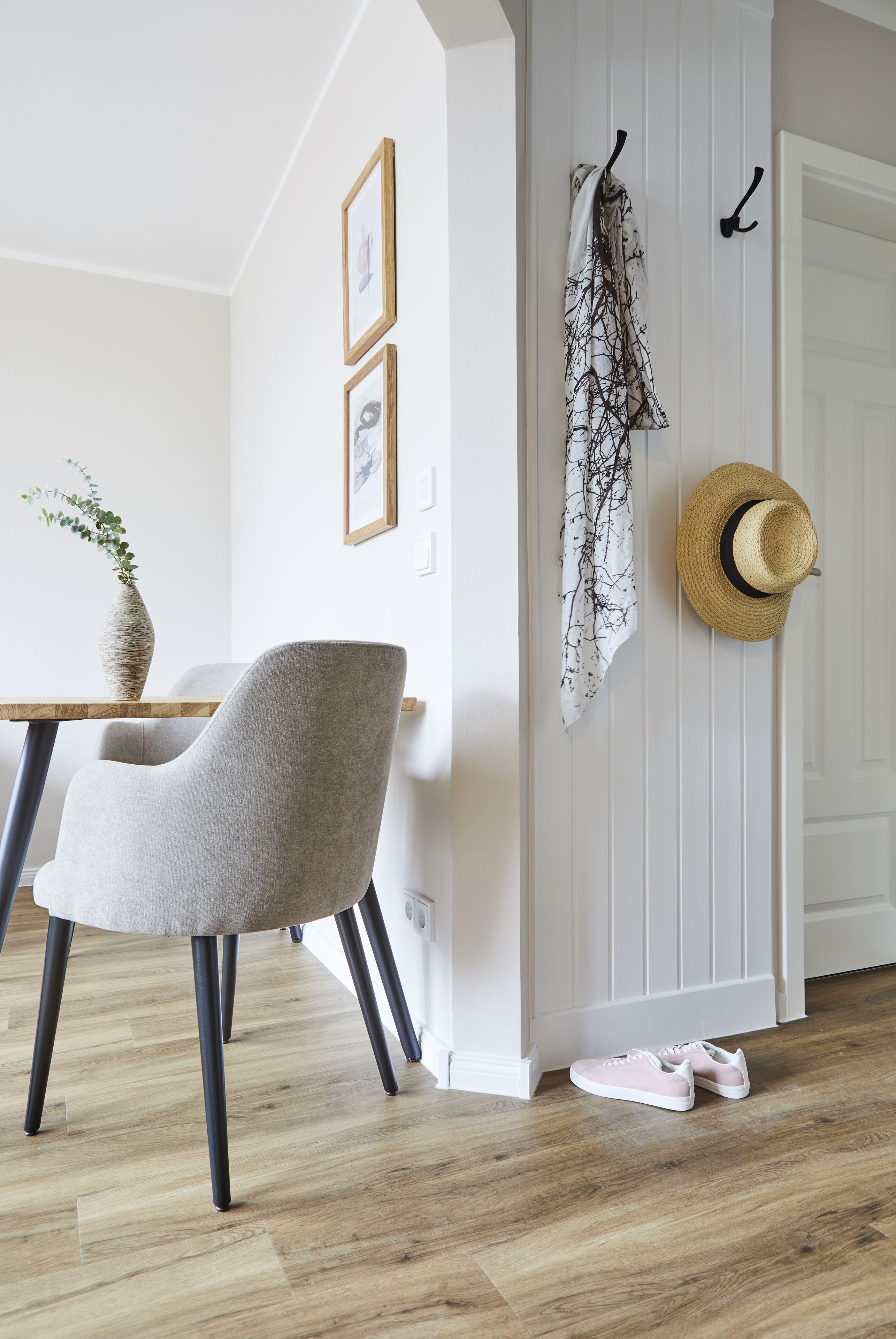 A room with a wooden dining table, a grey upholstered chair, a vase with a plant, a hat on the wall, and a pair of shoes on the floor.
