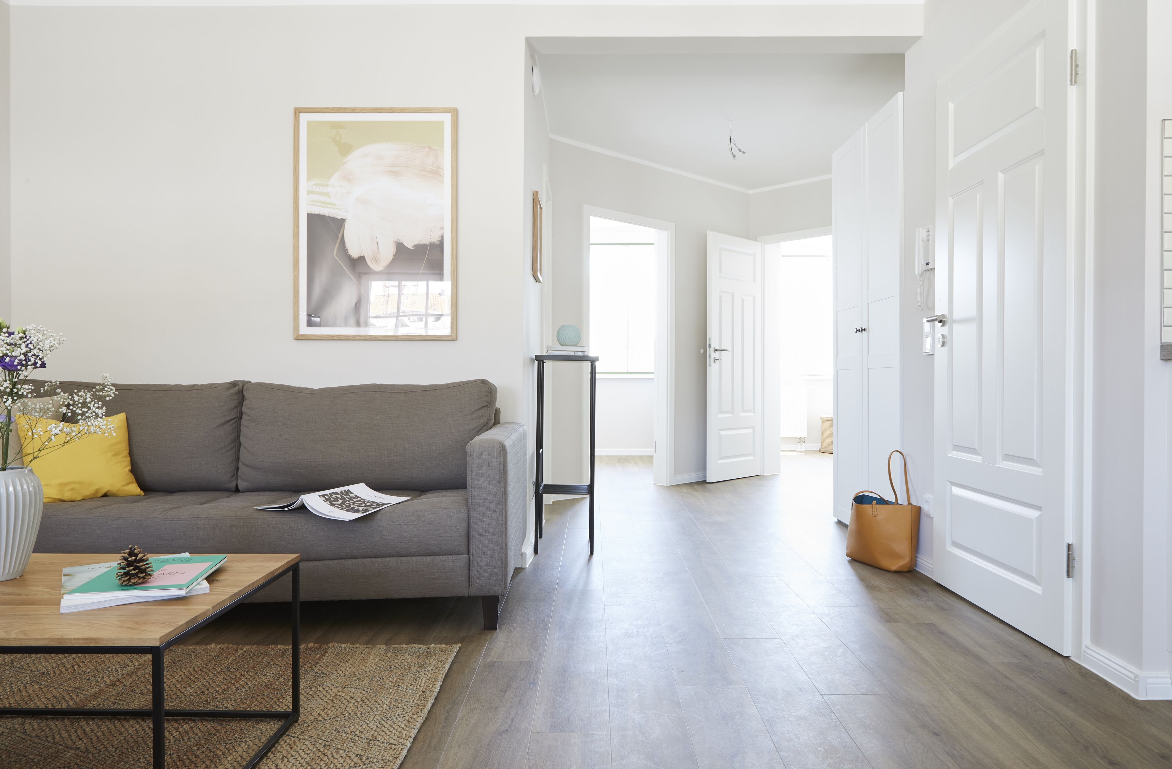 Un salón con un sofá gris, una mesa baja con un libro y un jarrón, un cuadro enmarcado en la pared, y un pasillo que conduce a una puerta.