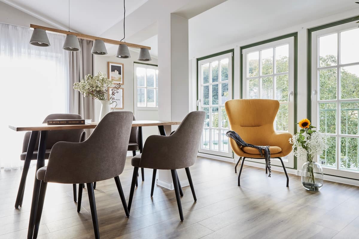 A modern dining area with a wooden table and grey chairs, large windows with green frames, and a yellow armchair.