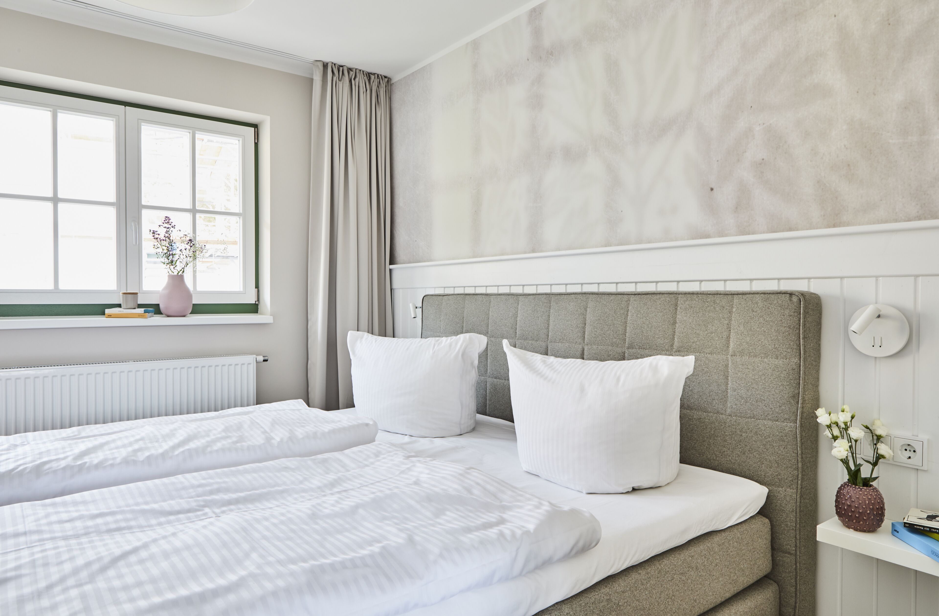 A modern hotel room with a large bed, a window with curtains, a radiator, and a wall-mounted light fixture.