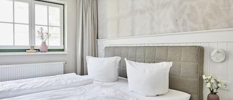 A modern hotel room with a large bed, a window with curtains, a radiator, and a wall-mounted light fixture.