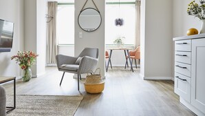 A modern living room with a grey armchair, a round mirror, a woven basket, and a dining area with orange chairs. - Villa Fortuna (Sellin)