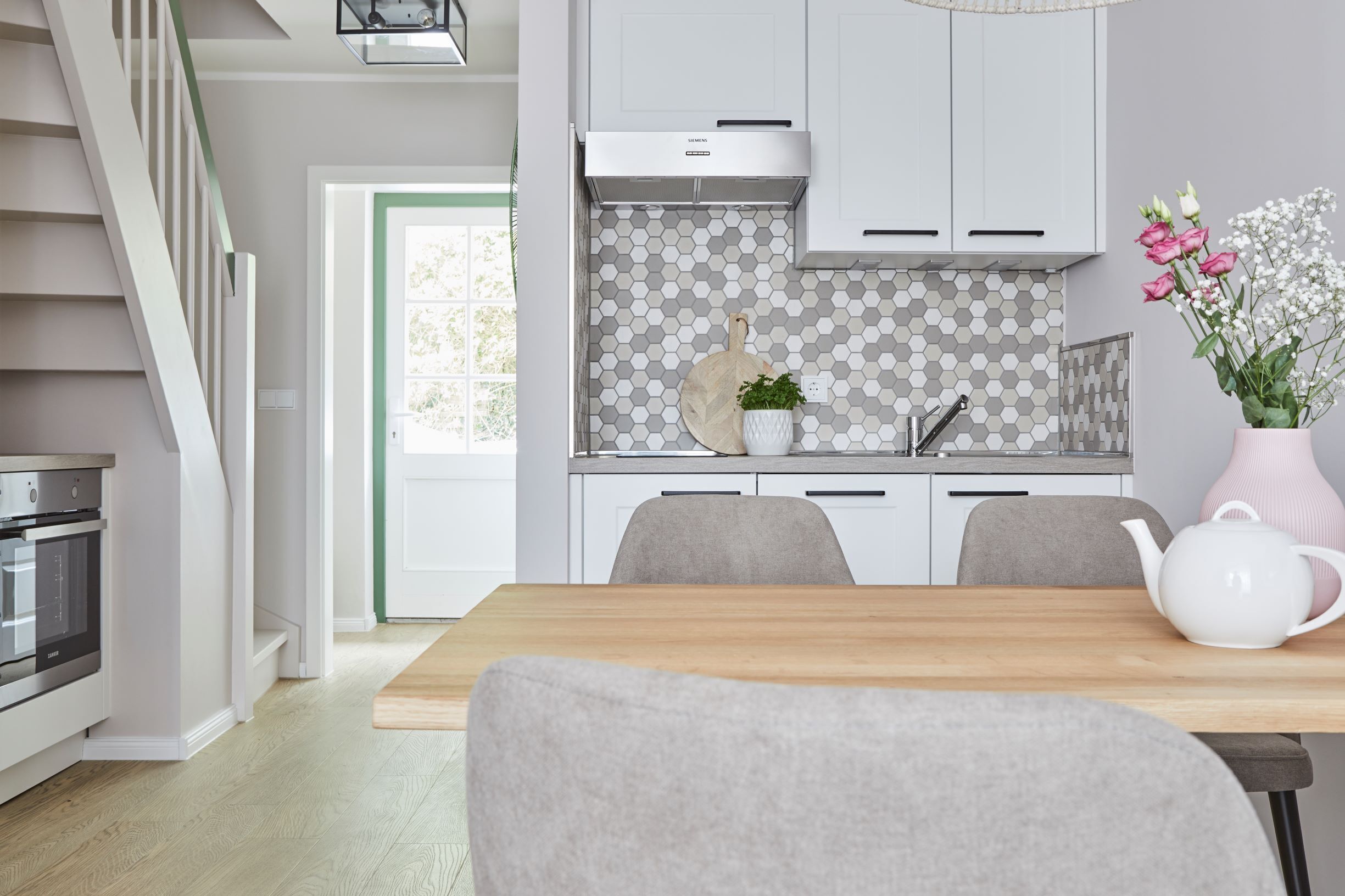 Uma cozinha moderna com armários brancos, painel hexagonal e uma área de refeições com mesa de madeira e cadeiras estofadas em cinza.