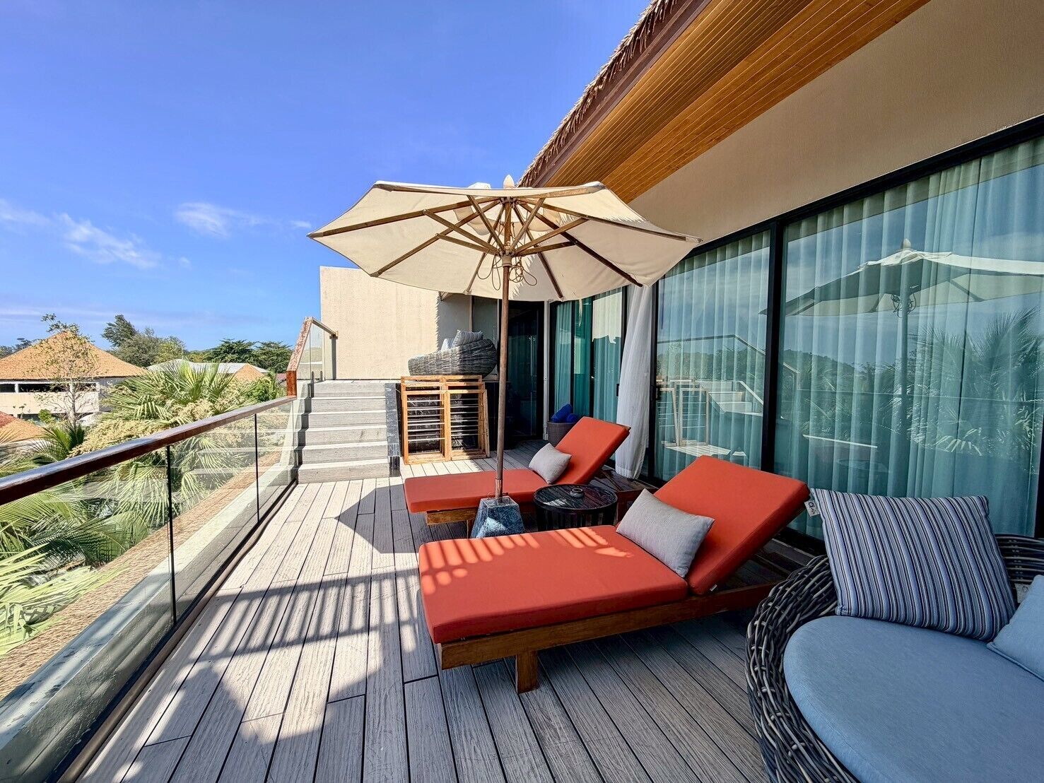A balcony with wooden flooring, red lounge chairs, a large umbrella, and a wicker chair.