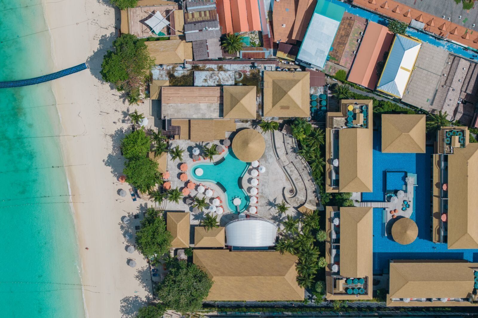 An aerial view of a resort with a central pool, multiple buildings, and a beach.