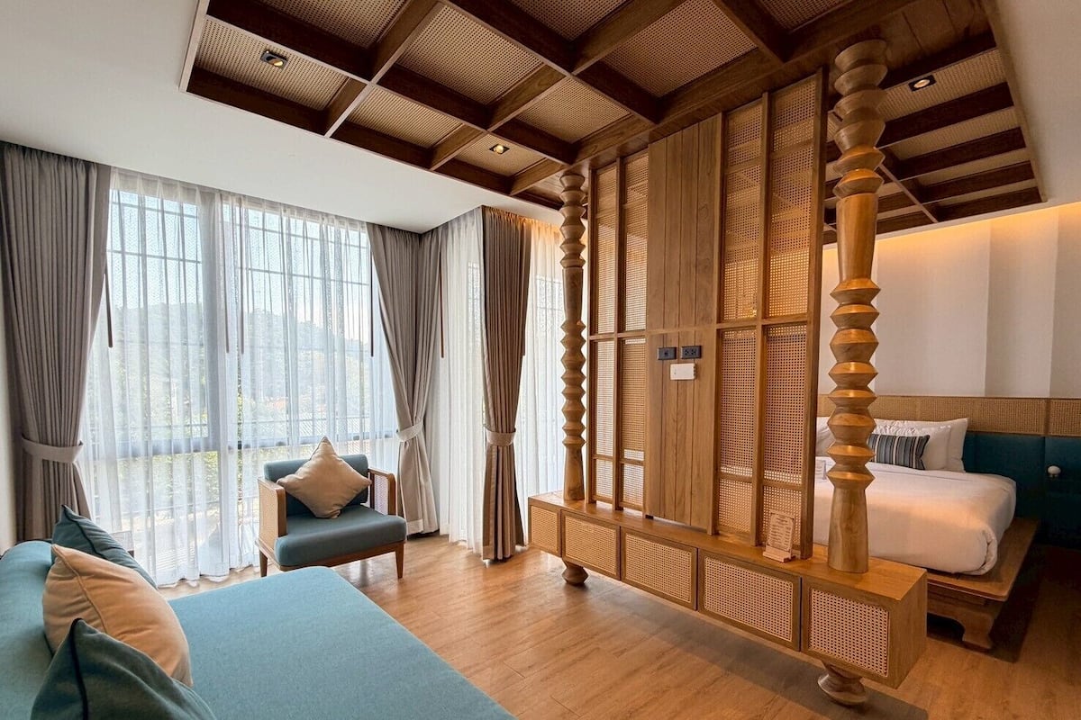 A hotel room with a large bed, a sofa, a wooden partition, and a view of the outdoors.
