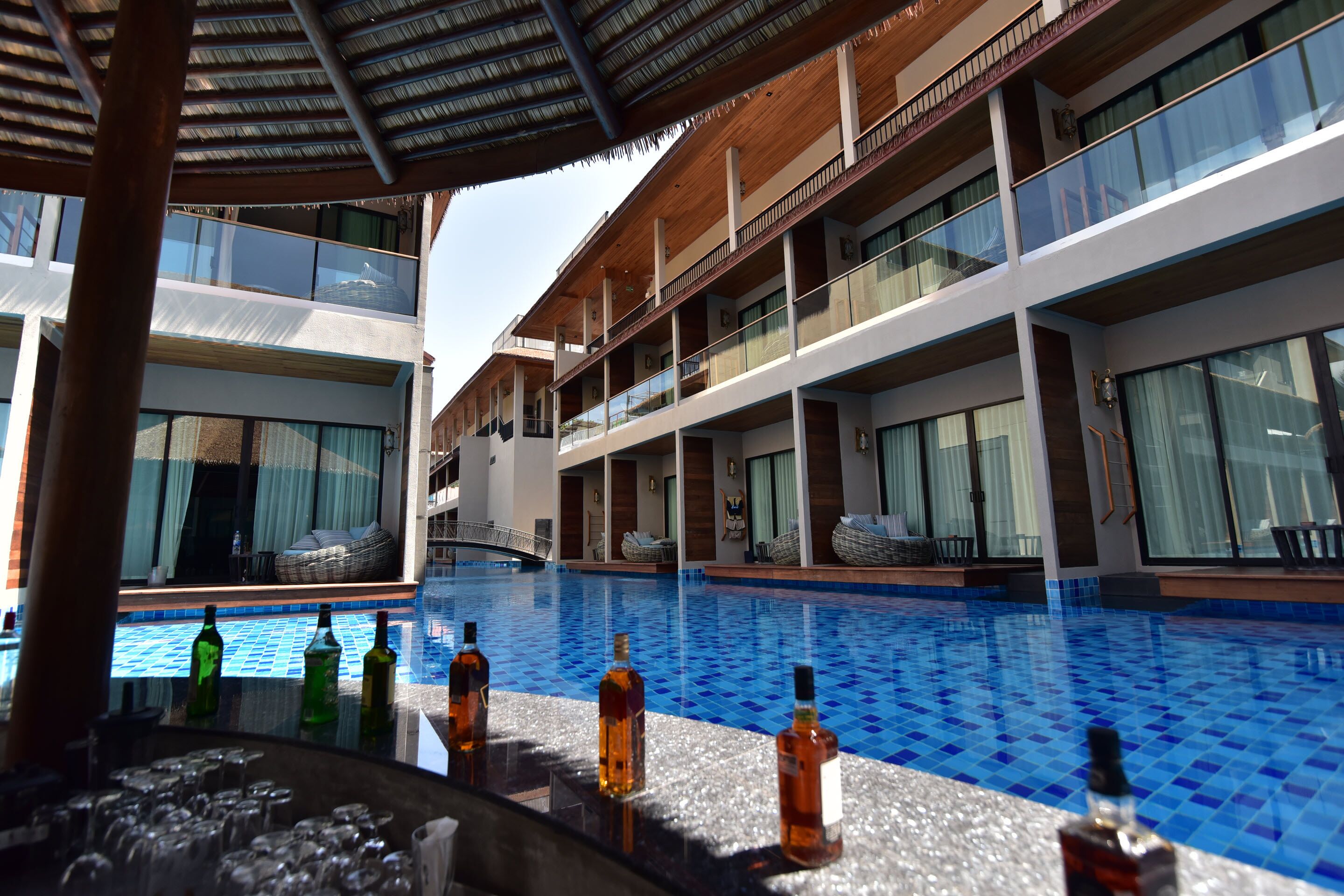 Un bar junto a la piscina con botellas y vasos, una piscina con azulejos azules y un edificio de varios pisos con balcones.