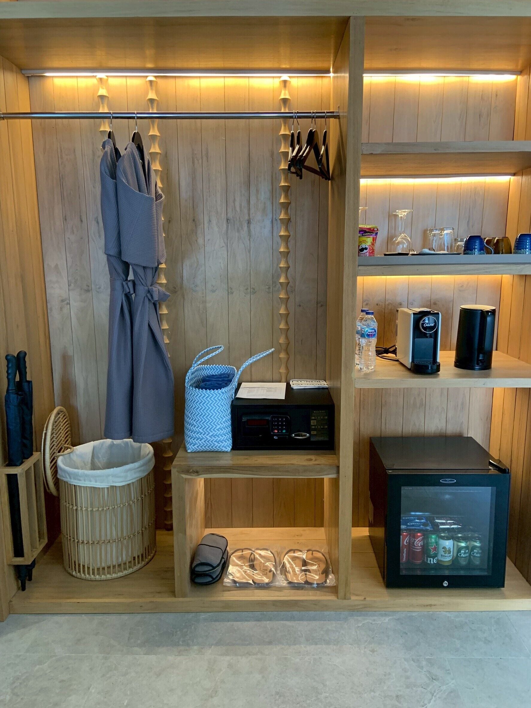 A compact room with a wooden interior, a small seating area with a chair, a trash bin, a microwave, a refrigerator, and a shelf with various items.
