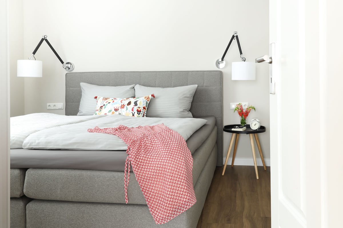 A neatly made bed with a grey headboard, two bedside lamps, and a small bedside table with a clock and a plant.