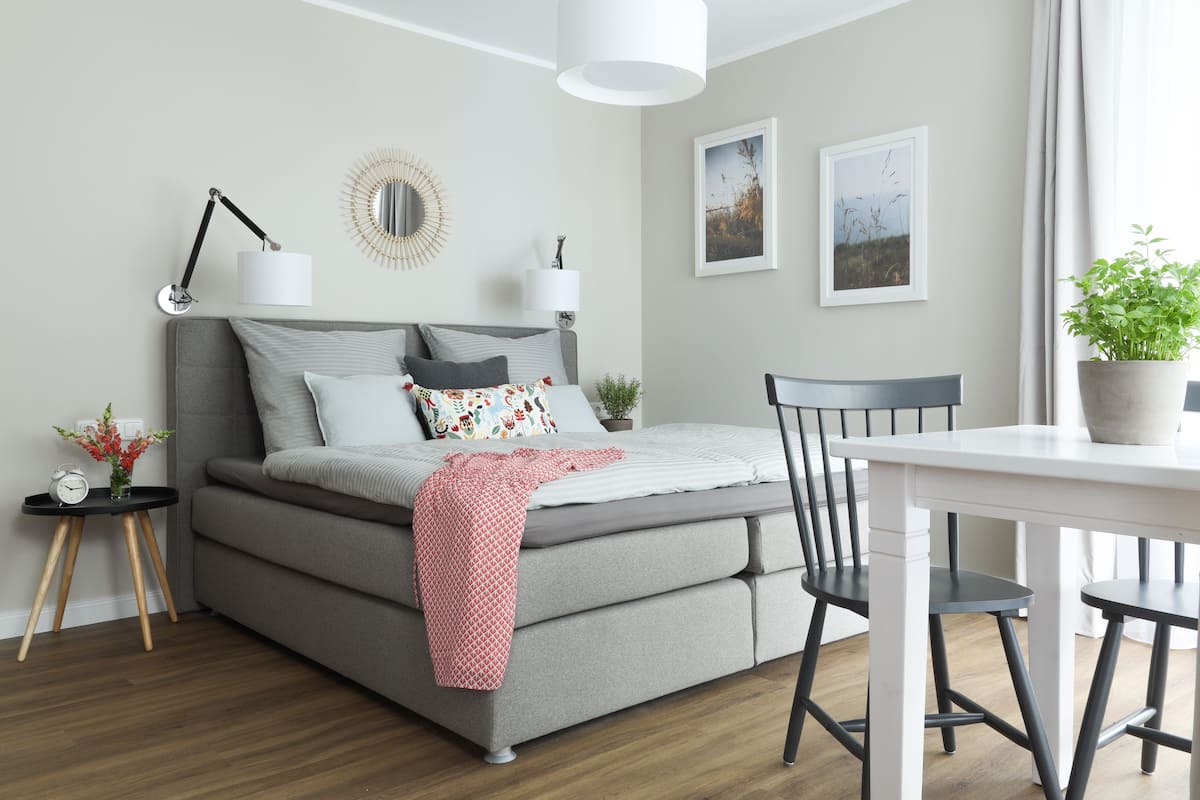 A modern bedroom with a grey sofa bed, a small bedside table with a lamp, a potted plant, and a dining table with two chairs.