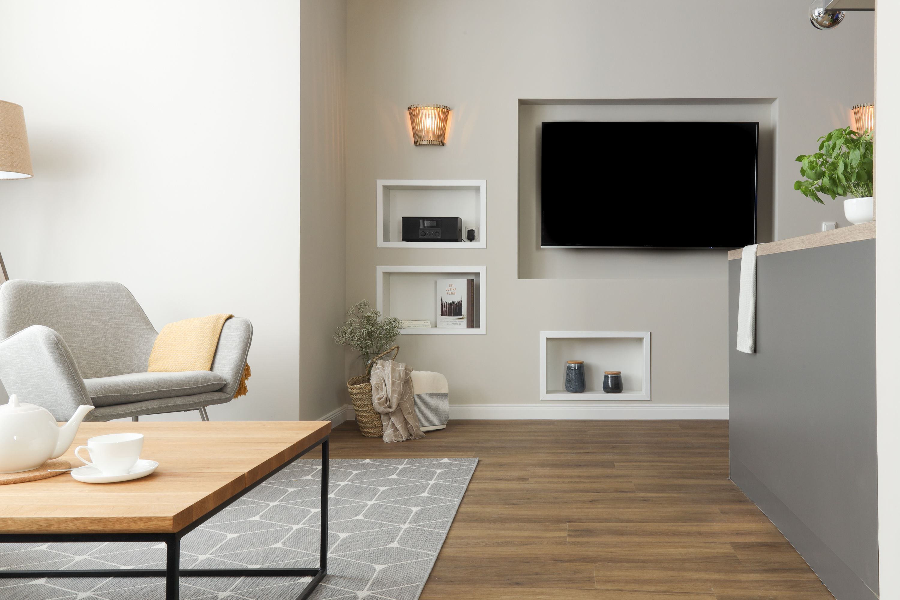 Uma sala de estar moderna com uma TV de tela plana na parede, um sofá cinza, uma mesa de centro de madeira com uma chaleira e xícara, e um tapete estampado.