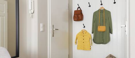 Un pasillo con un perchero que sostiene una camisa verde, una chaqueta amarilla y un bolso marrón. Una cesta de mimbre con una manta está en el suelo.