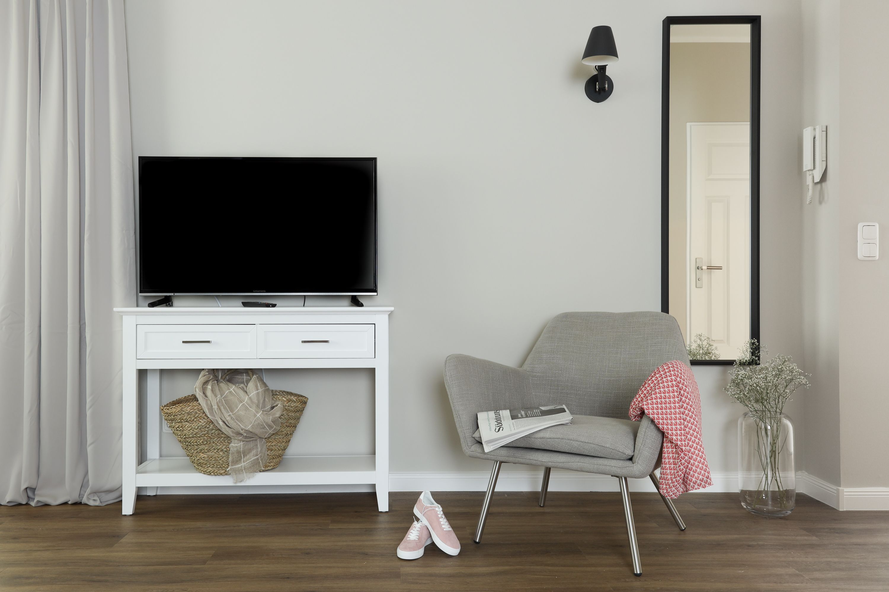 Ett modernt vardagsrum med en platt-TV på en vit TV-bänk, en grå fåtölj, ett litet bord med en lampa och en vas med blommor.