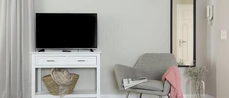 A modern living room with a flat-screen TV on a white console, a grey armchair, a small table with a lamp, and a vase with flowers.