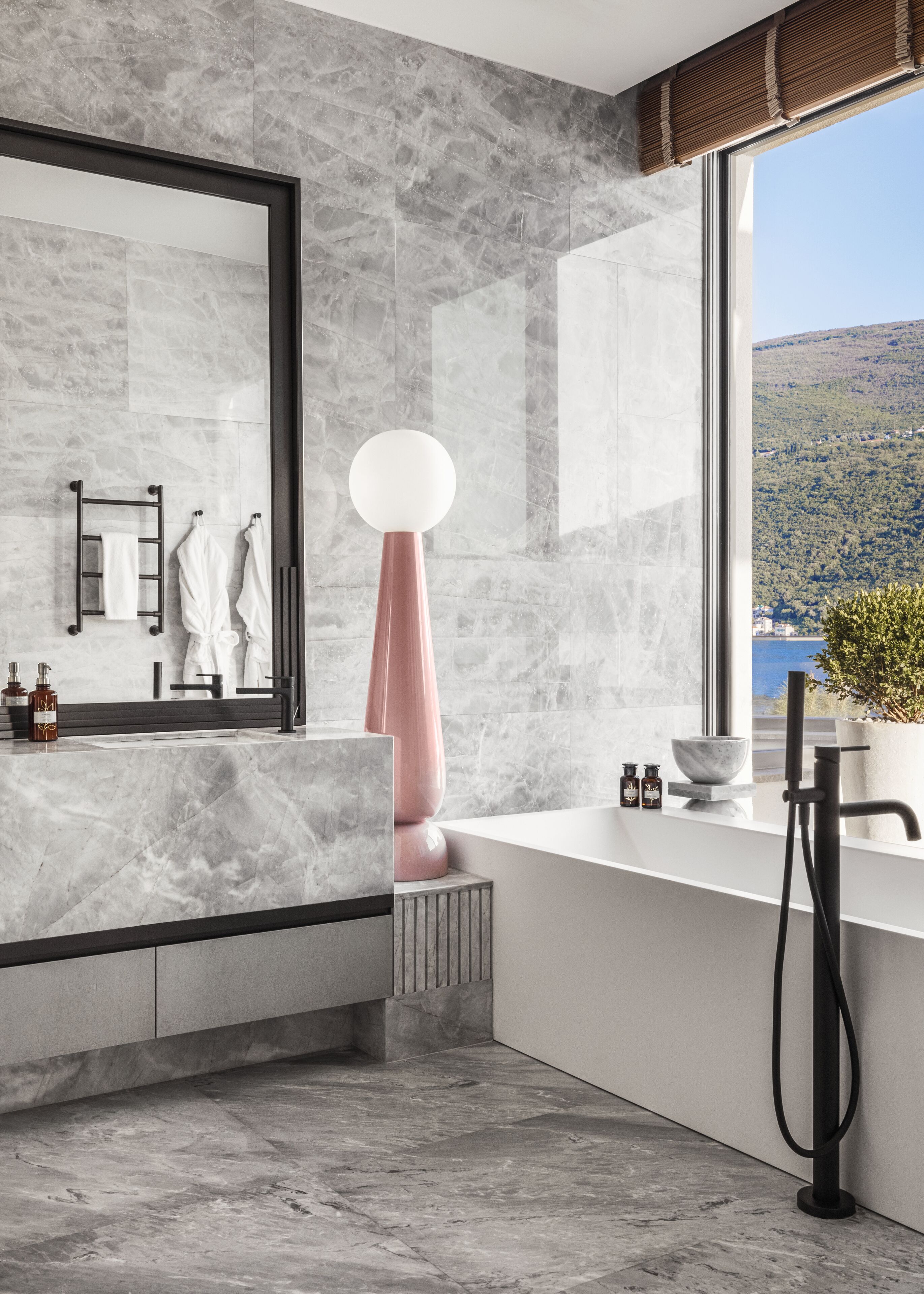 Ein modernes Badezimmer mit großem Spiegel, Marmorfliesen, einer weißen Badewanne und Blick auf einen See und Hügel.