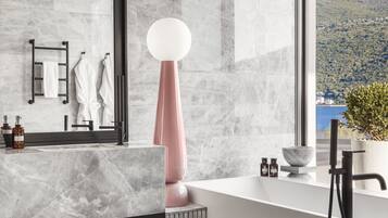 Ein modernes Badezimmer mit großem Spiegel, Marmorfliesen, einer weißen Badewanne und Blick auf einen See und Hügel.