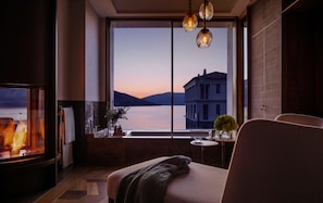 A cozy room with a fireplace, a comfortable sofa, a small table with a vase, and a large window overlooking a body of water and buildings. - One&Only Portonovi (Kumbor)