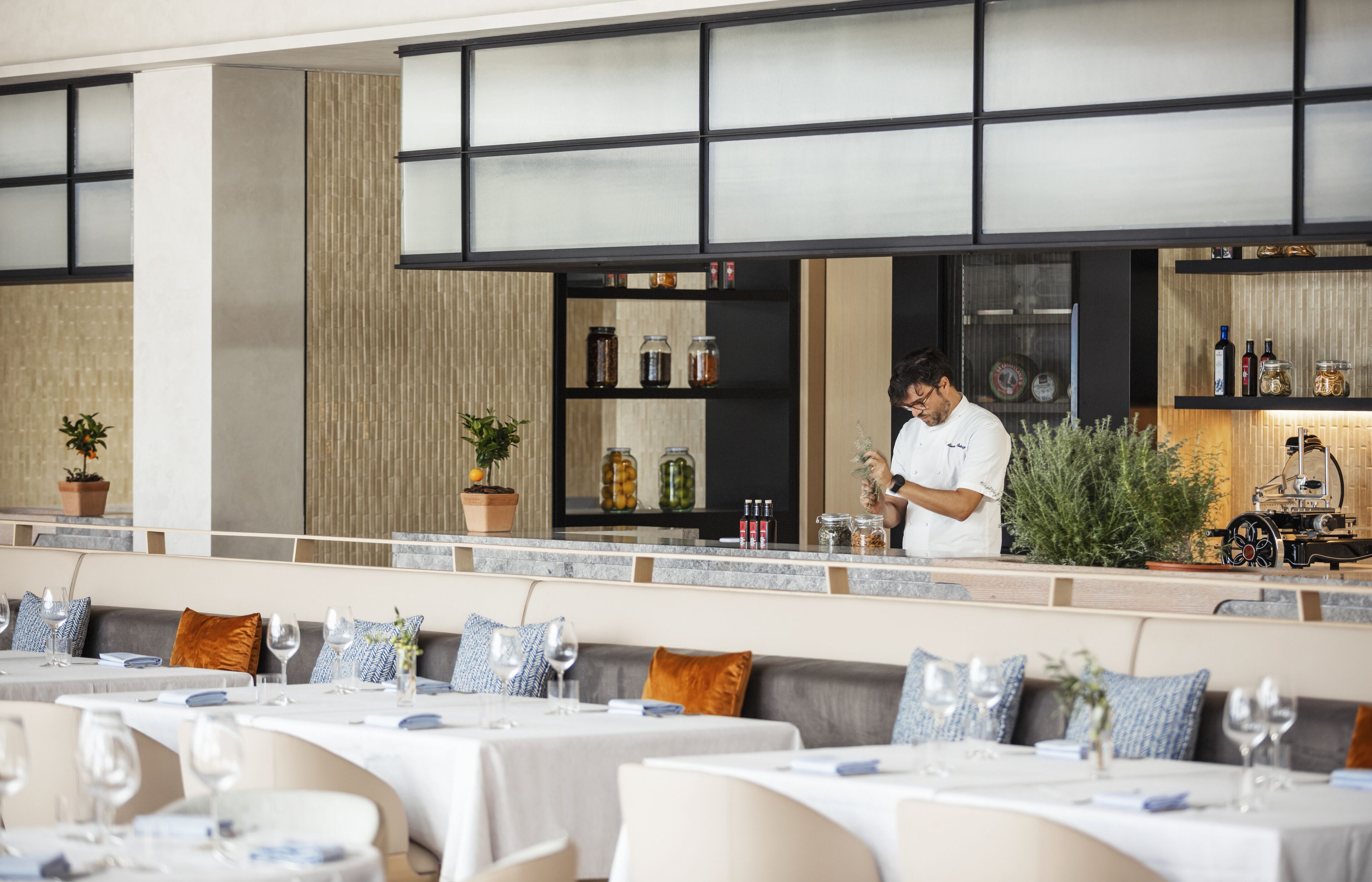 Un restaurante con un chef preparando comida en una barra, mesas con manteles blancos y servilletas azules, y estanterías con frascos y botellas al fondo.