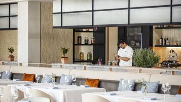 Un restaurante con un chef preparando comida en una barra, mesas con manteles blancos y servilletas azules, y estanterías con frascos y botellas al fondo.