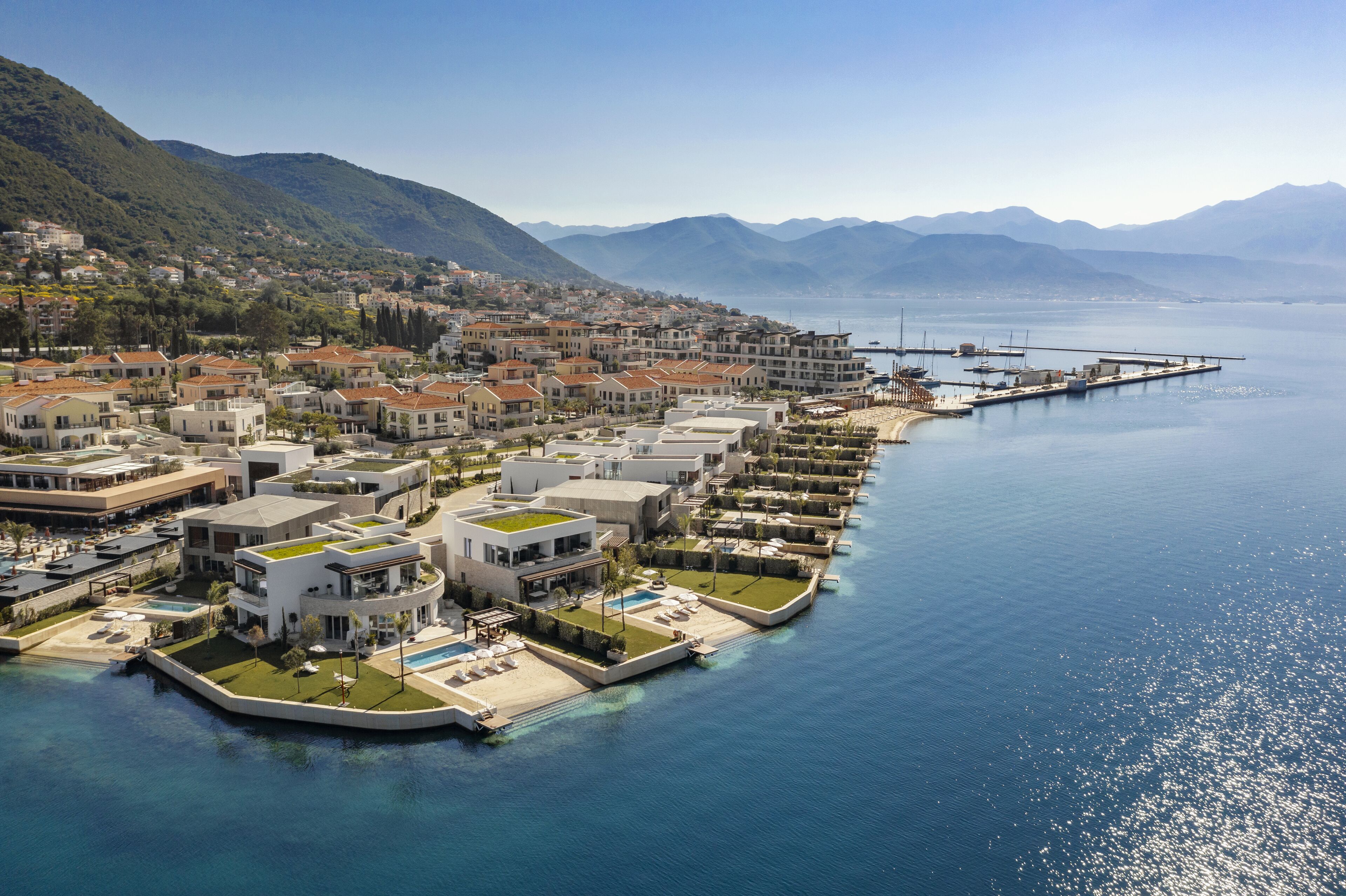 A coastal resort with modern architecture, a marina, and a mountainous backdrop.