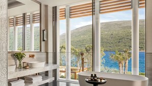 A modern bathroom with a freestanding bathtub, a large mirror, a vanity with a sink, and a view of a mountainous landscape through large windows. - One&Only Portonovi (Kumbor)