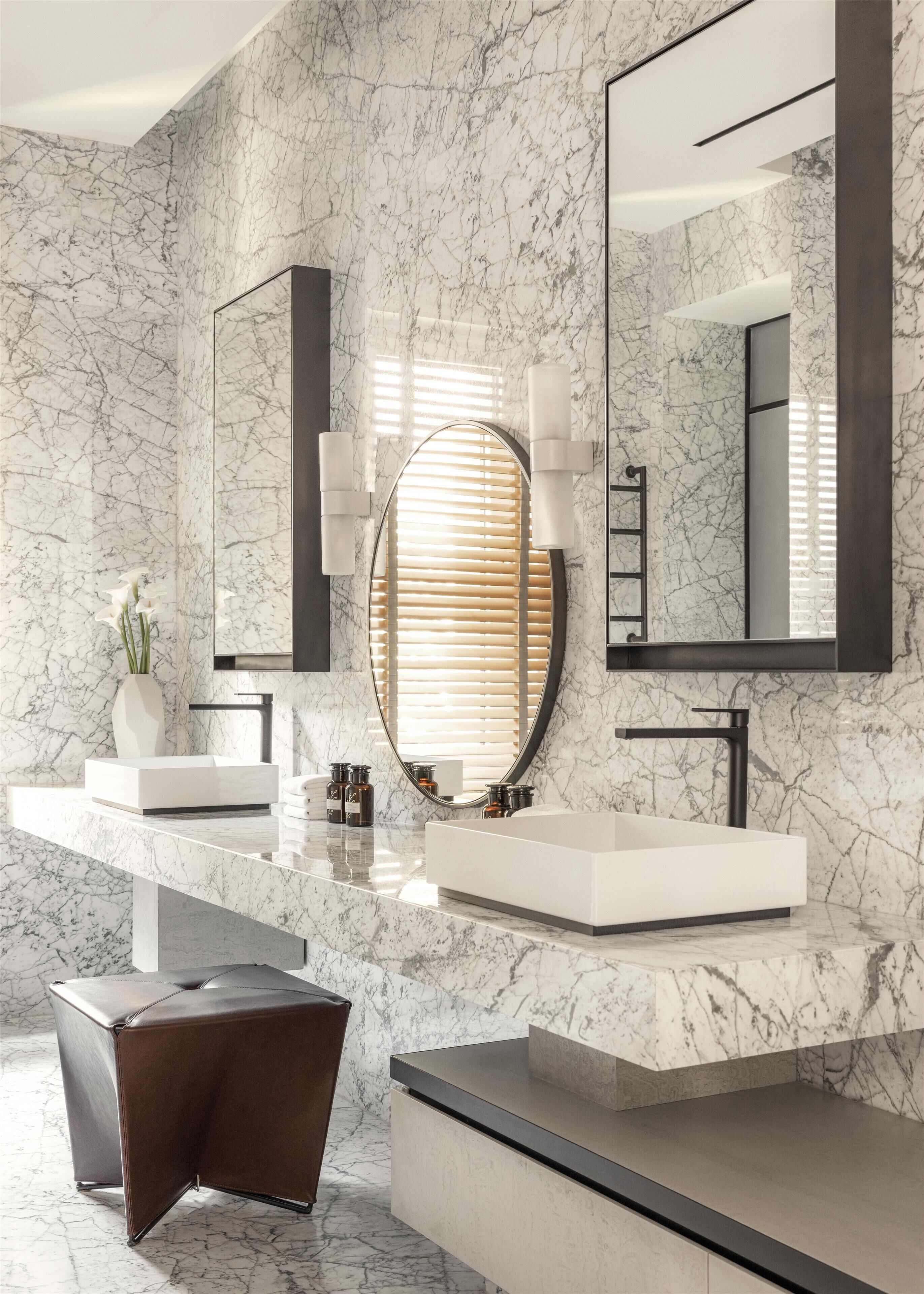 Un baño moderno con paredes de mármol, un espejo grande, dos lavamanos y un banco.
