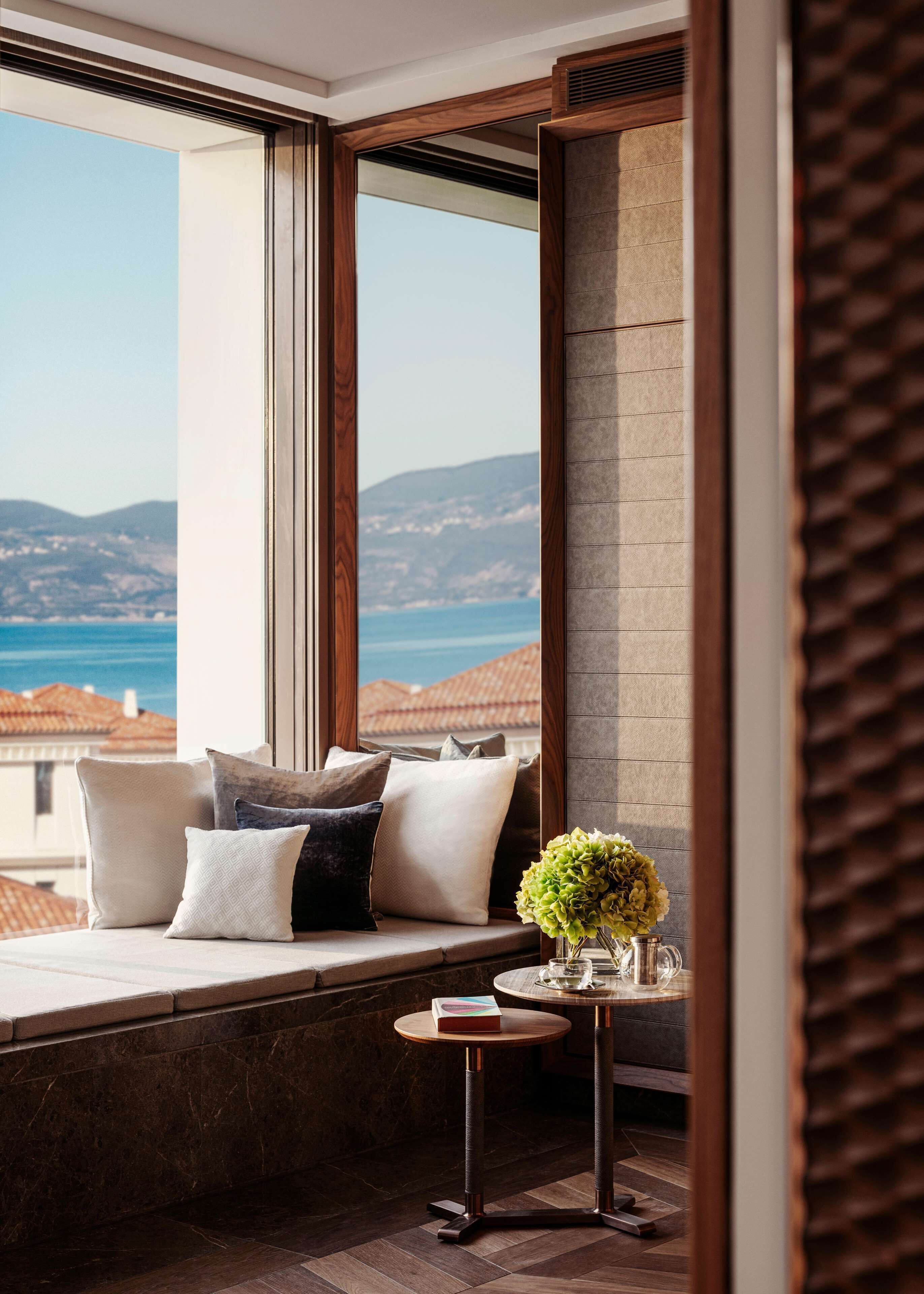 Una habitación con un ventanal grande, un banco acolchado, una mesita con un jarrón y vistas al mar y a las colinas.