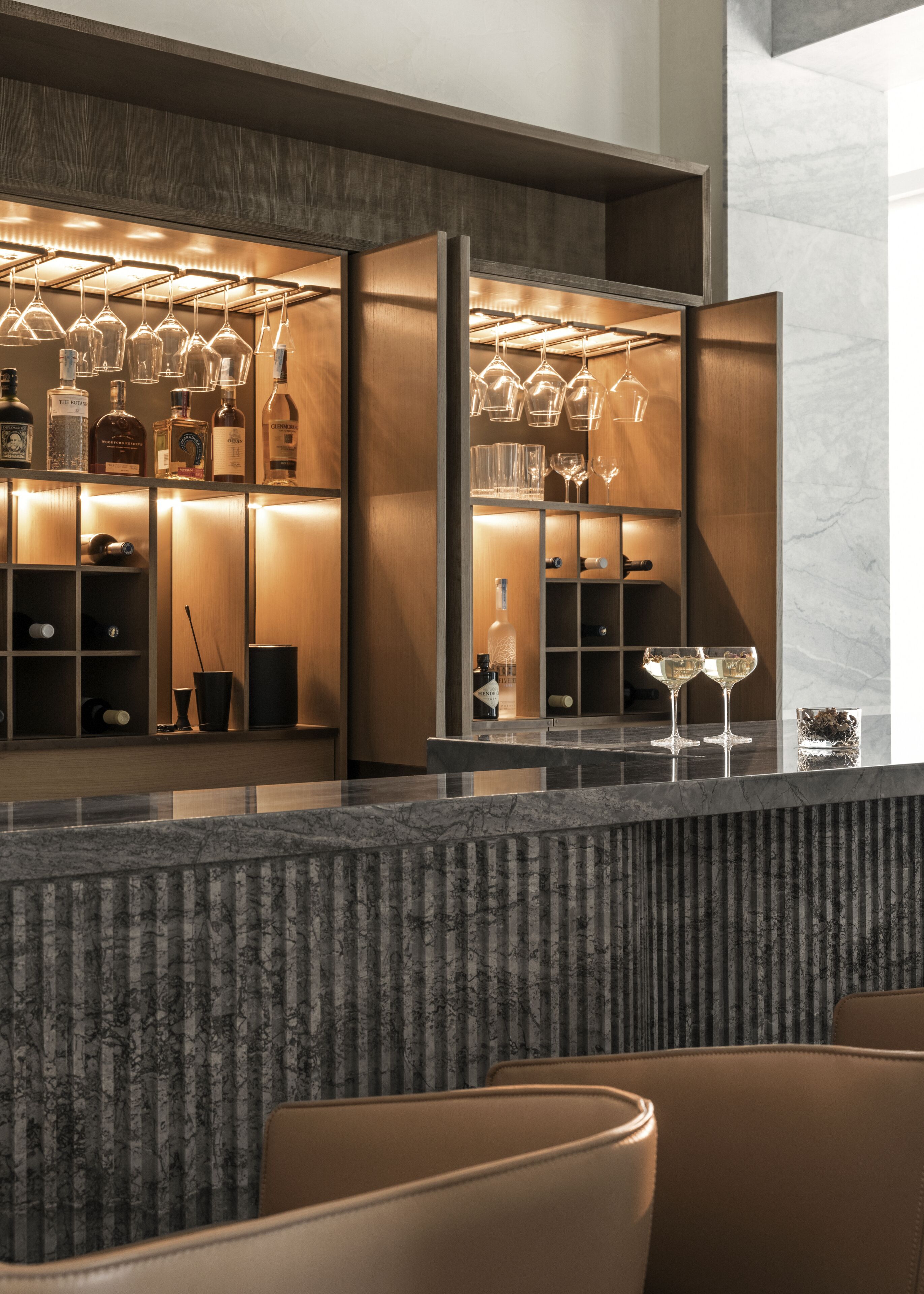 Una zona de bar moderna con estantes llenos de botellas y vasos, una encimera de mármol y sillones de cuero.