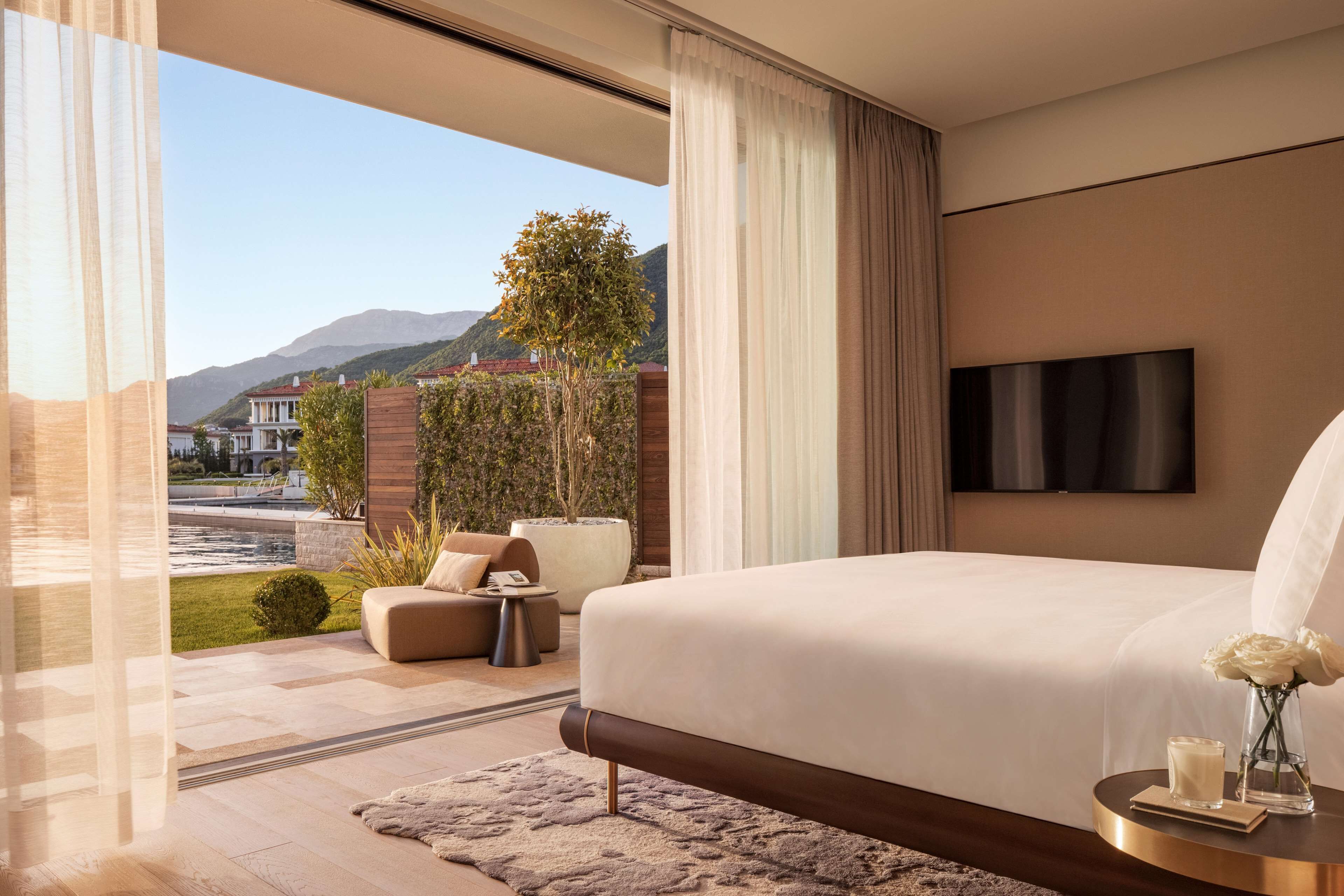 A modern bedroom with a large bed, a TV, and a view of a landscaped garden and mountains.