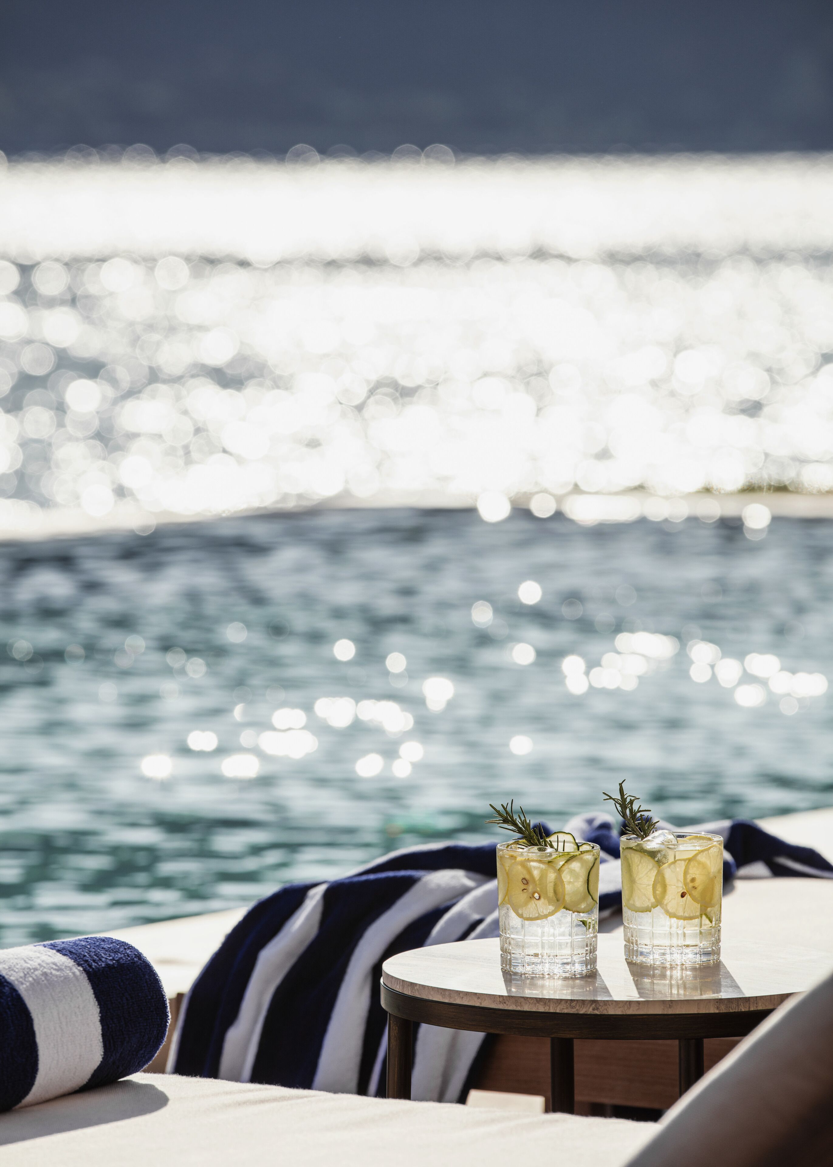 Two glasses of a beverage with lemon slices and a sprig of rosemary on a round table by a poolside with a view of the sea.