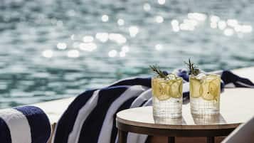 Two glasses of a beverage with lemon slices and a sprig of rosemary on a round table by a poolside with a view of the sea.