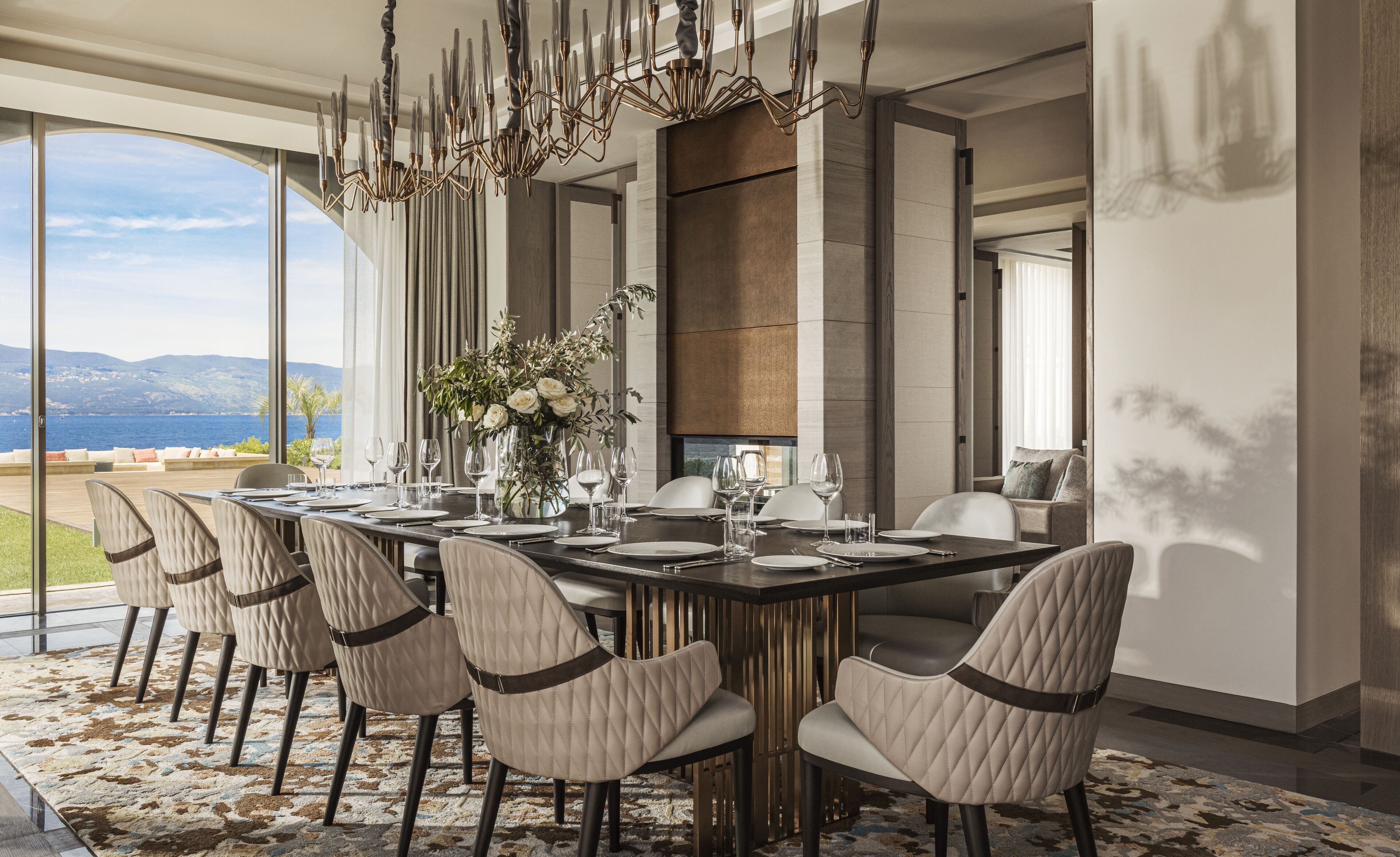 Une salle à manger moderne avec une longue table dressée pour six personnes, de grandes fenêtres offrant une vue sur la mer et un lustre décoratif.