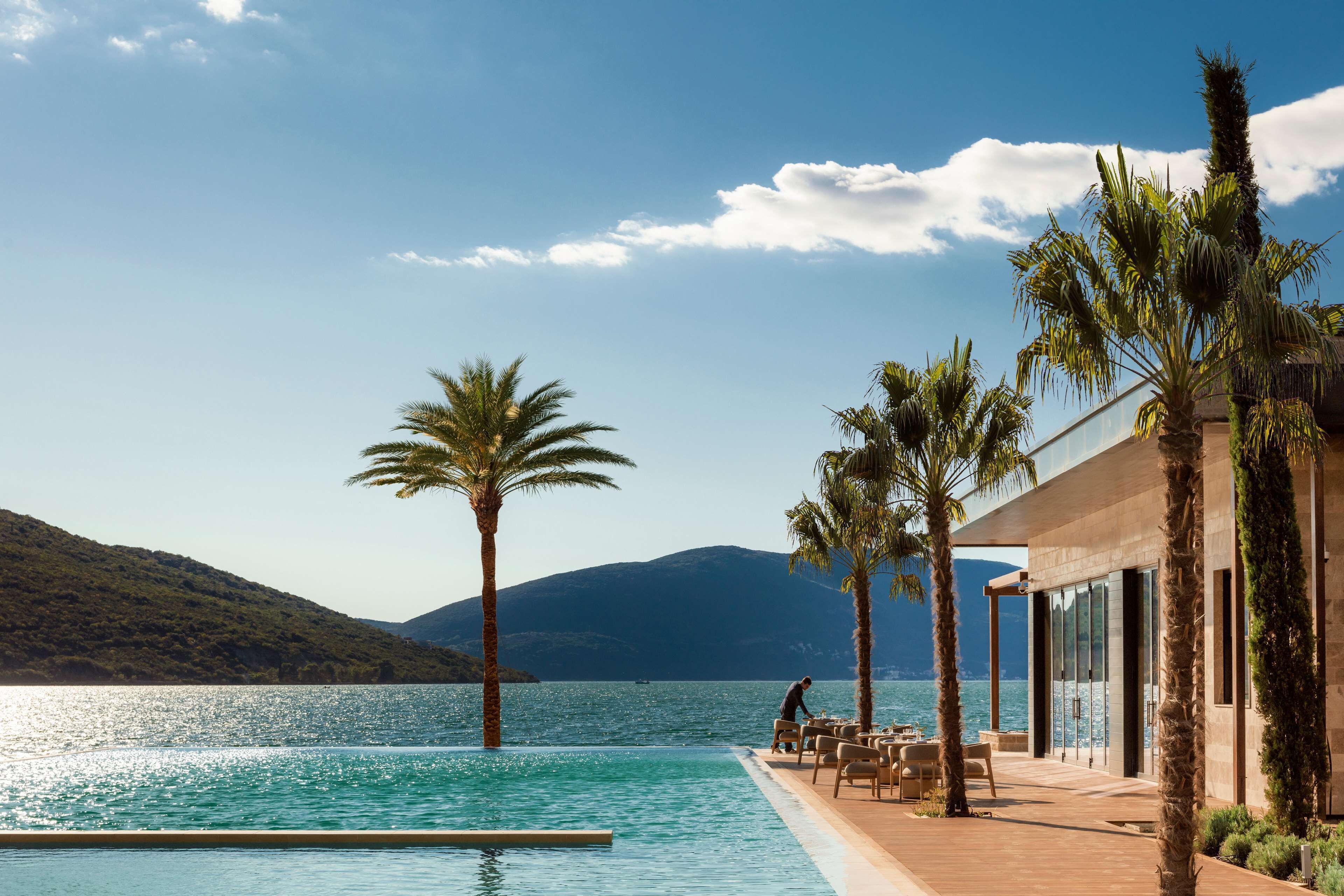 Una piscina con vistas al mar y a las montañas, palmeras y un edificio con amplios ventanales.