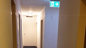 A narrow, carpeted hallway with white walls and doors on both sides, leading to a room with a visible exit sign. - Hotel Edelweiss (Neuhausen am Rheinfall)