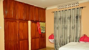 A bedroom with two beds, wooden wardrobes, a curtain with a pattern, and a wall decoration. - Dorabo Tourist Hotel (Jinja)