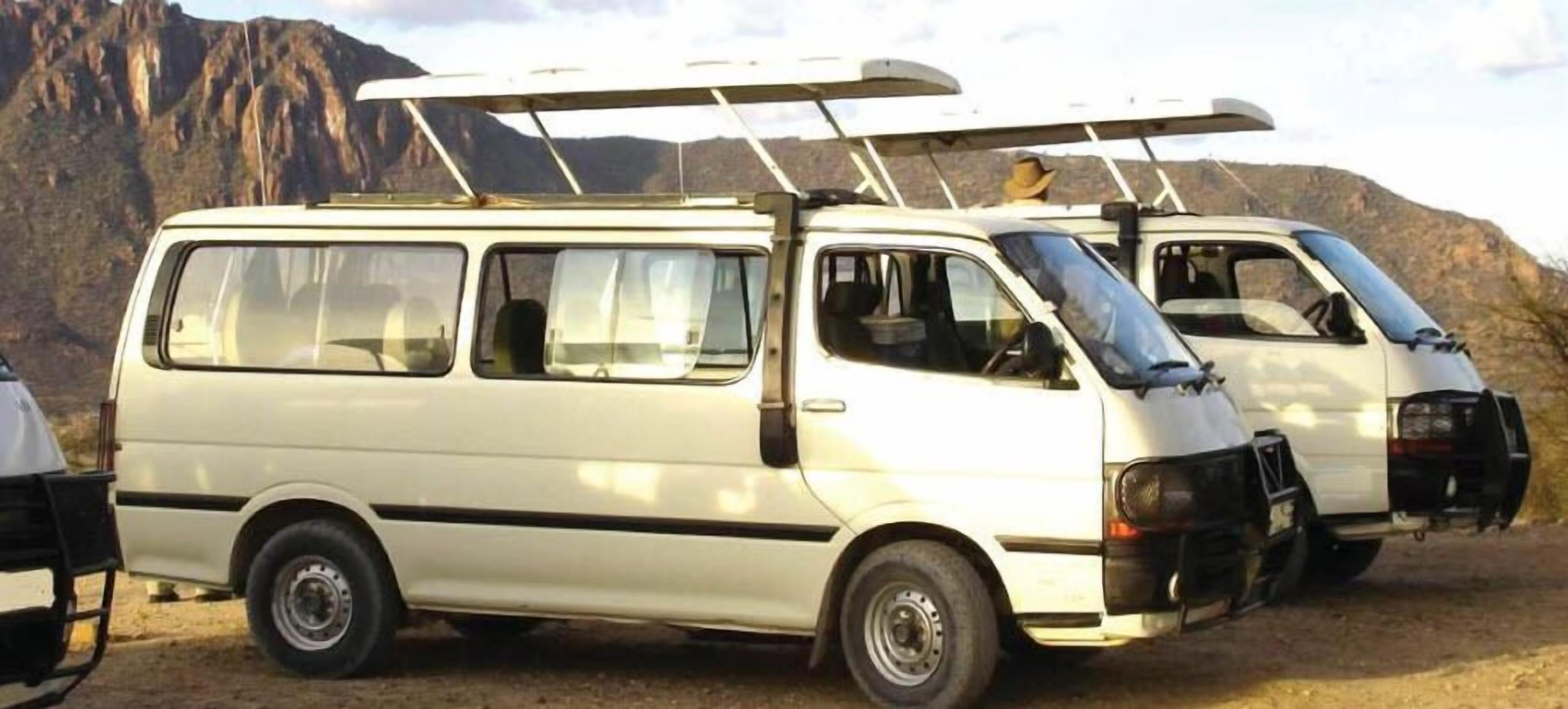 Una camioneta blanca con portaequipajes, estacionada al aire libre con montañas al fondo.