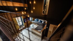 Una habitación estilo loft con cama en entrepiso, un pequeño rincón de descanso y un televisor fijado a una pared de ladrillo.