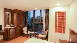 A hotel room with a bed, a desk with a mirror, a chair, a table with a cup, and a window with blinds.