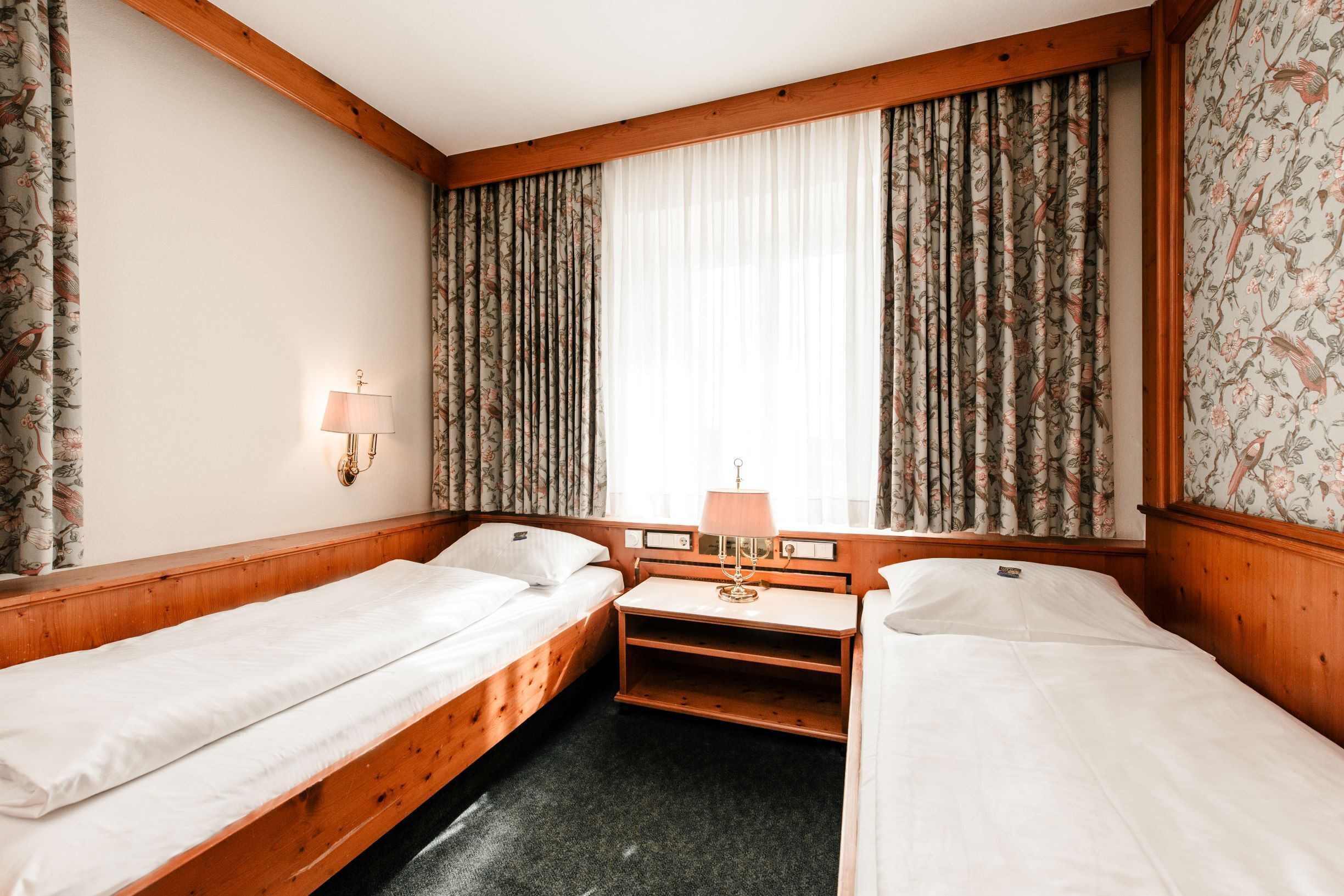 A room with two single beds, a small desk, a lamp, and a window with curtains.