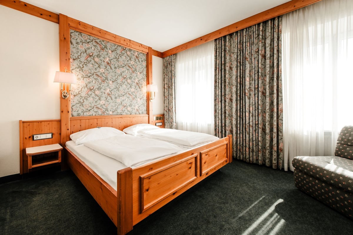 A wooden four-poster bed with a nightstand, a chair, and a large window with curtains.