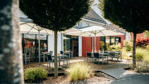 An outdoor seating area with tables and chairs under umbrellas, surrounded by greenery and a building with large windows. - Hotel Ochsen (Merklingen)