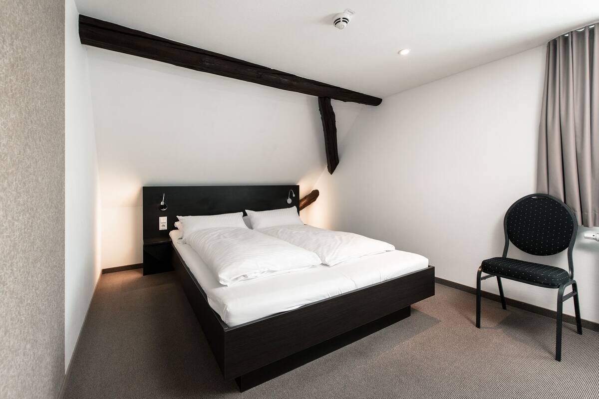 A modern hotel room with a dark wooden bed frame, white linens, a black upholstered chair, and a simple, clean design.